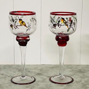May include: Two clear glass candle holders with a red rim and base. Each holder features a painted bird and floral design. The stem is clear, and the base is round. The candle holders are decorative and suitable for home decor.