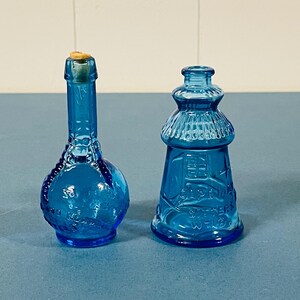 May include: Two cobalt blue glass bottles. The left bottle has a round base, a long neck, and a cork stopper. The right bottle is shaped like a lighthouse with a wide base, a tapered body, and a narrow neck, featuring embossed text and patterns.