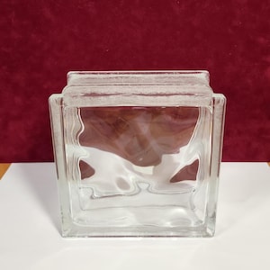 May include: A clear, square glass block with a textured, wavy interior design. The block is transparent, allowing light to pass through. The edges are clean and defined, creating a modern aesthetic. The block is suitable for construction or decorative purposes.
