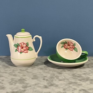May include: A vintage tea set featuring a white teapot and a teacup with saucer. The teapot and teacup are decorated with pink and red floral designs and green accents. The set is on a gray surface with a blue background.