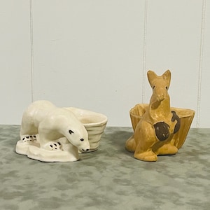 May include: Two ceramic animal figurines. One is a white polar bear with a cub, and the other is a tan kangaroo with a pouch. Both have a basket-like container attached.