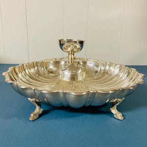 May include: A silver-toned metal serving dish with a scalloped, shell-like design and a central raised bowl. The dish features a polished finish, a wide rim with decorative edges, and stands on three ornate, claw-shaped feet for stability.