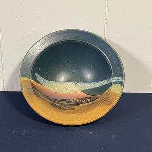 May include: A round ceramic bowl with a landscape design. The bowl features a dark blue interior, a light blue and white band, and a tan exterior. The landscape design includes pink, brown, and tan elements.