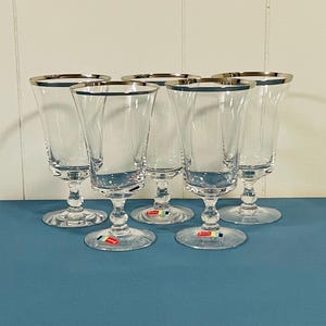 May include: Five clear stemmed glasses with silver rims are arranged on a blue surface. The glasses have a classic design with a flared bowl and a slender stem. The background is a white wall.
