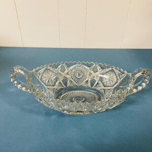 May include: A clear pressed glass oval bowl featuring a complex geometric starburst pattern, a serrated sawtooth rim, and two curved handles on opposite sides. The base of the bowl displays a detailed cross-hatched design with a central star motif.