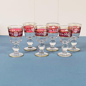 May include: Set of six clear glass cordial glasses. Each glass features a red band with white floral designs and gold trim. The glasses have a round base and stem, and are arranged on a blue surface.