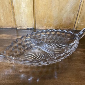 May include: Clear glass serving dish with a leaf shape. The dish has a central divider and a textured surface with a scalloped edge. The dish is designed for serving snacks or appetizers.