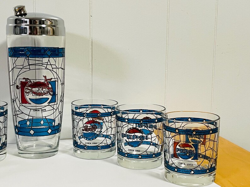 May include: A vintage Pepsi cocktail shaker and glasses set. The clear glass pieces feature a blue and white stained-glass design with the Pepsi logo and the text "Circa" followed by a year.