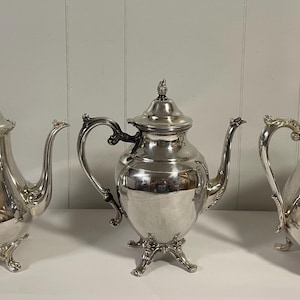 May include: A silver tea set including a teapot, a coffee pot, and a creamer. The set has ornate handles and feet. The teapot and coffee pot have lids with decorative finials. The creamer has a curved spout.