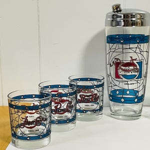 May include: A vintage Pepsi-Cola cocktail set featuring a cocktail shaker and three glasses. The glasses and shaker have a blue and white geometric design with the Pepsi logo and the text "Circa" with dates. The shaker has a silver lid.