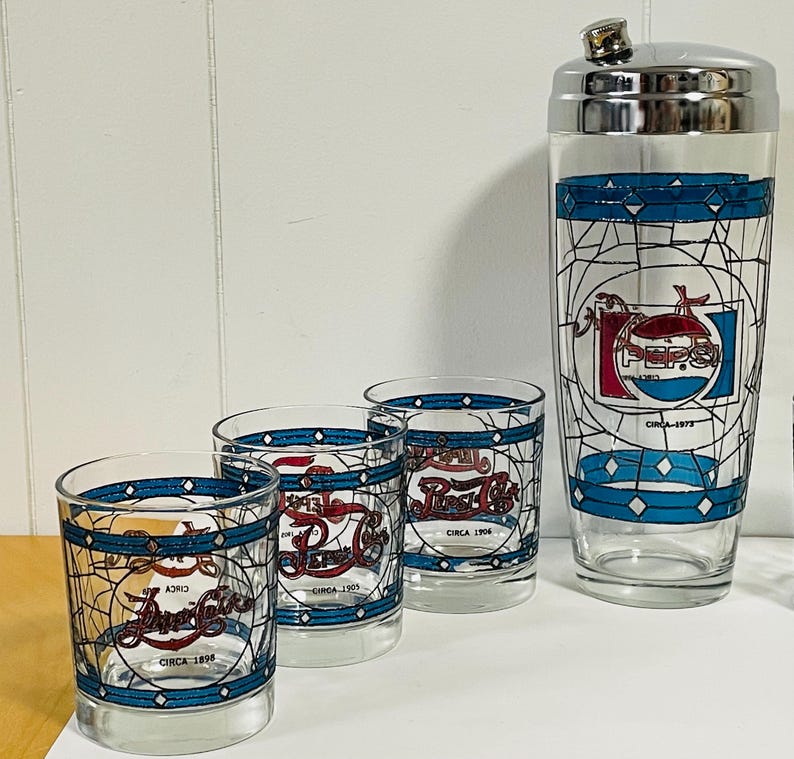 May include: A vintage Pepsi-Cola cocktail set featuring a cocktail shaker and three glasses. The glasses and shaker have a blue and white geometric design with the Pepsi logo and the text "Circa" with dates. The shaker has a silver lid.