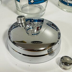 May include: A shiny chrome flask with a screw-on cap and a separate, smaller cap. The flask's surface reflects the surrounding objects. The image also shows several clear drinking glasses with a blue and white design.