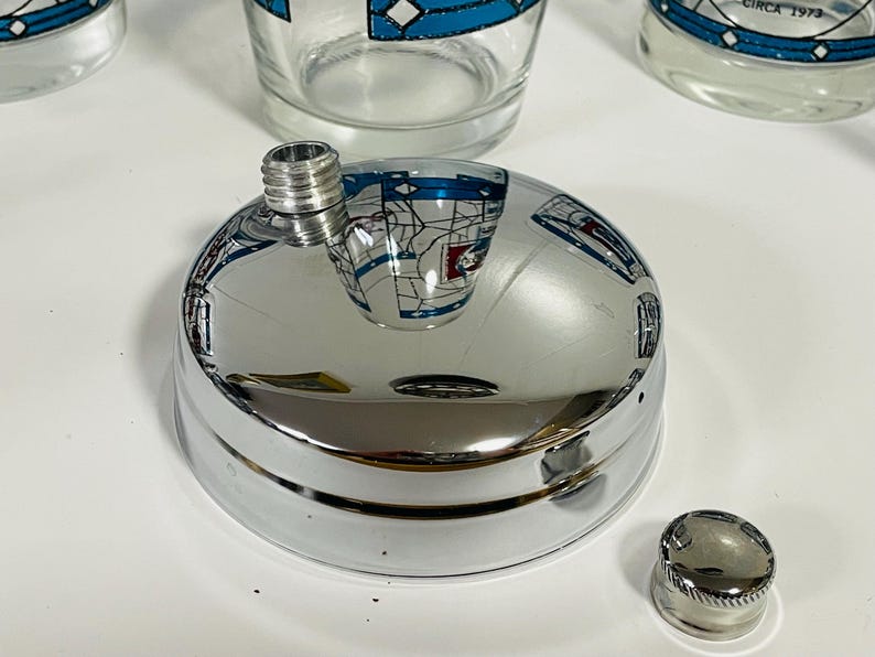 May include: A shiny chrome flask with a screw-on cap and a separate, smaller cap. The flask's surface reflects the surrounding objects. The image also shows several clear drinking glasses with a blue and white design.