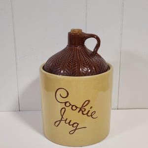 May include: A vintage cookie jug with a brown lid and handle, and a tan base. The lid has a textured pattern. The words "Cookie Jug" are written in a cursive font on the base. The jug is made of ceramic.