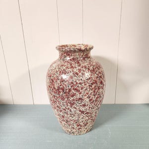 May include: A ceramic vase featuring a speckled pattern with a mix of deep red, maroon, and black spots against a cream-colored base. The vessel has a rounded body, a tapered base, and a flared rim, showcasing a glossy finish throughout the exterior.