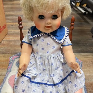 May include: A vintage doll with blonde hair, wearing a blue and white dress, sitting on a heart-shaped patterned cushion.