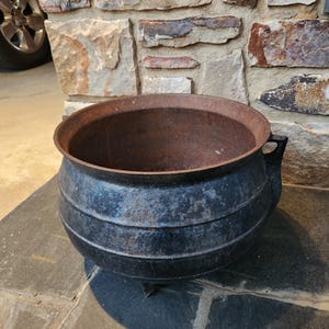May include: A heavy, round cast iron cauldron with a dark black exterior and a rusted, weathered interior. The pot features a wide, open top, a rounded belly with horizontal ridges, and three short, sturdy legs for stability on flat surfaces.