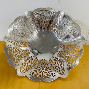 May include: A silver-colored, decorative bowl with an intricate, openwork design. The bowl has a scalloped edge and a smooth, reflective interior. The openwork pattern features a floral or leaf-like motif, adding a touch of elegance to the piece.