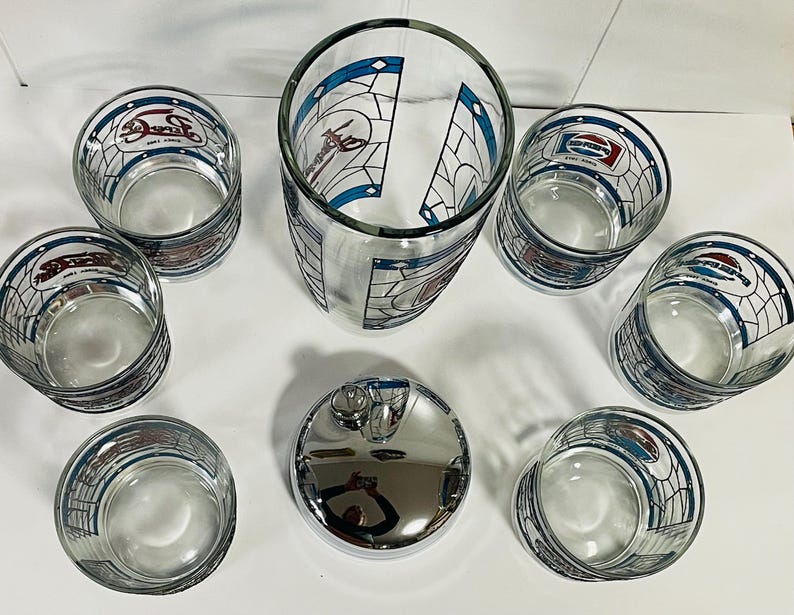 May include: A set of clear glass Pepsi-branded drinking glasses and a chrome-finished bottle stopper. The glasses feature a stained-glass design with the Pepsi logo and are arranged around a tall glass and the stopper.