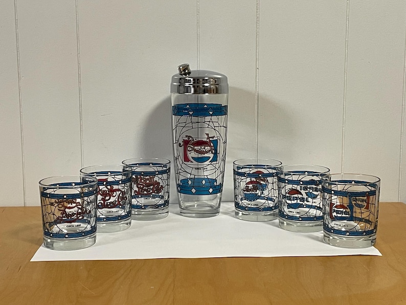 May include: A vintage Pepsi-Cola cocktail set featuring a shaker and six glasses. The glasses and shaker have a blue and white design with the Pepsi logo and the text "Since 1893". The set is displayed on a white surface.