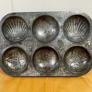 May include: A vintage metal baking pan with six individual molds. The molds have different designs, including shells and leaves.