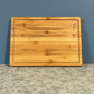 May include: A rectangular bamboo cutting board with a juice groove around the edge. The board has a natural wood grain pattern and is set against a blue background.