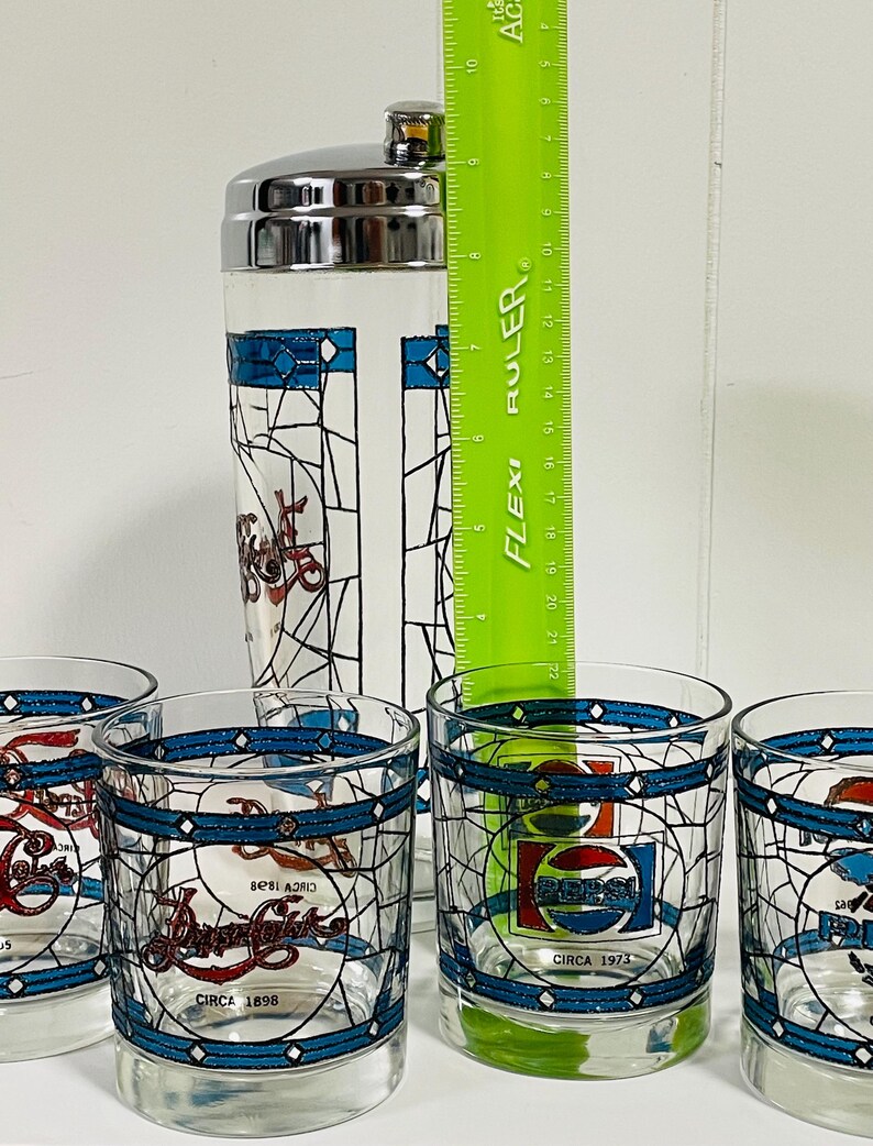 May include: A vintage Pepsi-Cola cocktail shaker and glasses set. The clear glass shaker has a silver lid and a stained-glass design. The glasses feature the Pepsi logo and the text "CIRCA 1898" and "CIRCA 1973". The ruler shows the shaker is approximately 8 inches tall.