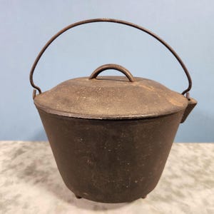 May include: A vintage cast iron Dutch oven with a lid and handle. The pot is dark brown with a slightly textured surface and three small feet. The handle is a curved metal bar. The lid has a small handle.