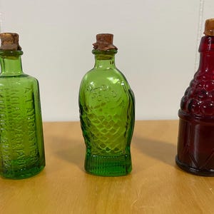 May include: Three small glass bottles with cork stoppers. The bottles are green and red. The green bottles have a textured surface. The red bottle has a design of grapes.