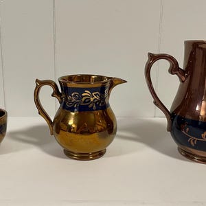 May include: Three copper-colored ceramic pitchers with blue and gold accents. The largest pitcher has a wide mouth and a handle. The two smaller pitchers have a narrower mouth and a handle. All three pitchers have a floral design.