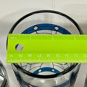 May include: Clear glass with a black rim and a blue and white geometric design. A green ruler is placed across the top of the glass, measuring approximately 3 inches in diameter.