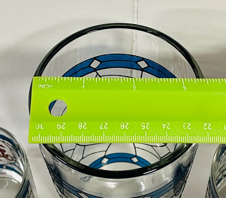 May include: Clear glass with a black rim and a blue and white geometric design. A green ruler is placed across the top of the glass, measuring approximately 3 inches in diameter.