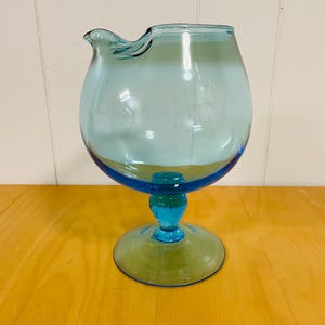 May include: A vintage, light blue glass pitcher with a rounded body and a spout. The pitcher sits on a circular base, supported by a short stem. The glass has a gradient effect, transitioning from a darker blue at the base to a lighter blue at the top.