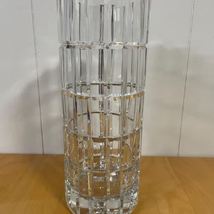 May include: A clear, cylindrical crystal vase with a geometric, textured design. The vase features vertical and horizontal cuts, creating a grid-like pattern. The vase is approximately 10 inches tall and is suitable for displaying flowers or as a decorative piece.