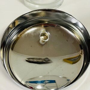 May include: Close-up of a shiny, chrome-colored metal lid with a small, circular handle in the center. The lid has a slightly concave shape and reflects the surrounding environment. The image also shows a clear glass with a blue and white geometric design.