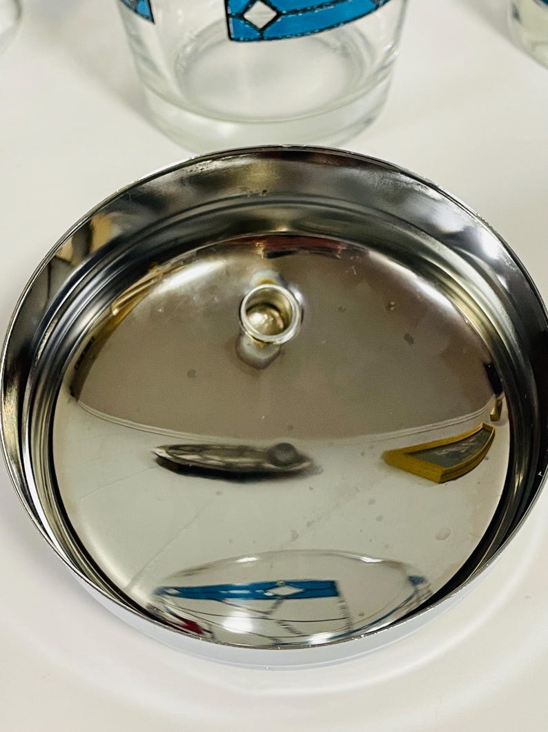 May include: Close-up of a shiny, chrome-colored metal lid with a small, circular handle in the center. The lid has a slightly concave shape and reflects the surrounding environment. The image also shows a clear glass with a blue and white geometric design.