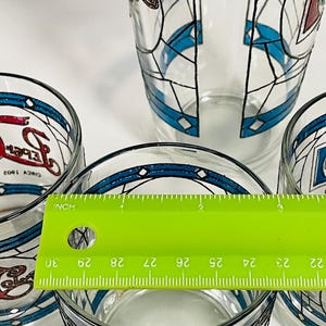 May include: A set of clear glass tumblers with a stained-glass design. The glasses feature blue and red accents with black outlines. A green ruler shows the diameter of the glass is approximately 2.5 inches.