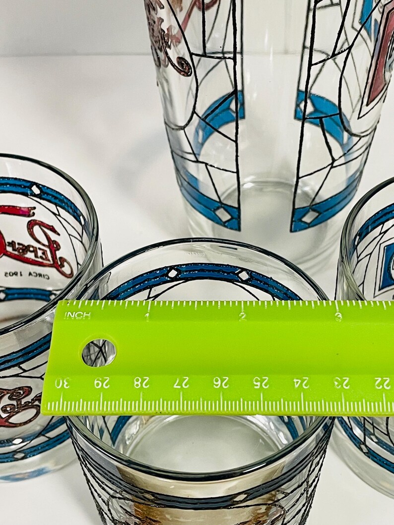 May include: A set of clear glass tumblers with a stained-glass design. The glasses feature blue and red accents with black outlines. A green ruler shows the diameter of the glass is approximately 2.5 inches.