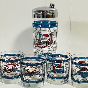 May include: A vintage Pepsi-Cola cocktail shaker and six matching glasses. The clear glass pieces feature the Pepsi logo in red, white, and blue, with a blue geometric border. The shaker has a silver lid and the text "Circa 1906" is visible.