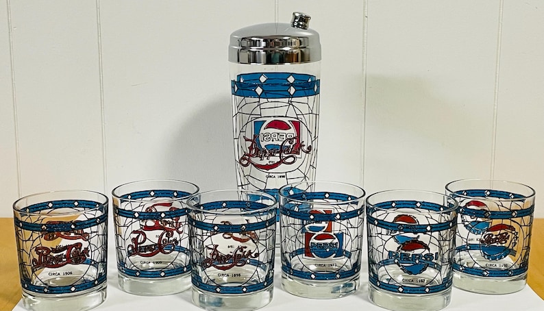 May include: A vintage Pepsi-Cola cocktail shaker and six matching glasses. The clear glass pieces feature the Pepsi logo in red, white, and blue, with a blue geometric border. The shaker has a silver lid and the text "Circa 1906" is visible.