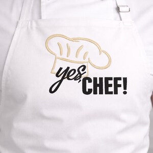 Może przedstawiać: Biały fartuch z haftowaną złotą czapką szefa kuchni i napisem "yes, CHEF!" w kolorze czarnym. Fartuch jest noszony na białym płaszczu kucharskim. Prosty, odważny design dla entuzjastów gotowania.