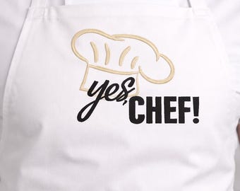 May include: White apron with a gold embroidered chef's hat and the words "yes, CHEF!" in black script. The apron is worn over a white chef's coat. The design is simple and bold, perfect for culinary enthusiasts.