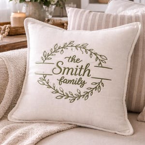 May include: Cream-colored square throw pillow with the embroidered text "the Smith family" in green. The text is surrounded by a green leafy wreath design. The pillow is on a beige sofa with a matching throw.