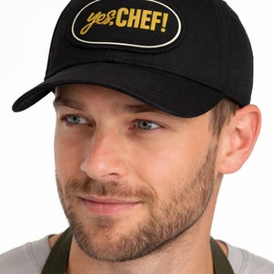 May include: Black baseball cap with a curved peak. The cap has a gold and white oval patch with the words "yes chef!" in a script font. The cap is worn by a person.