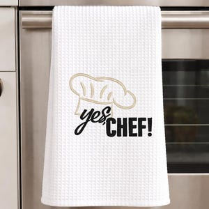 Può includere: Canovaccio da cucina bianco a trama a nido d'ape con la scritta "yes, CHEF!" in corsivo nero e lettere maiuscole. Un cappello da chef è ricamato in beige chiaro sopra il testo. L'asciugamano è appeso a un forno in acciaio inossidabile.