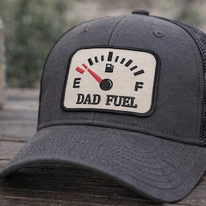 May include: A charcoal gray trucker hat with a mesh back. The front patch features a fuel gauge design with a red needle pointing to empty and the words "DAD FUEL". A can is visible in the background.