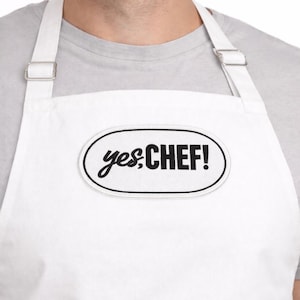 Può includere: Grembiule bianco con spalline regolabili. Il grembiule presenta la scritta "yes, CHEF!" in corsivo nero all'interno di un ovale bordato di nero. Il grembiule è progettato per l'uso in cucina.