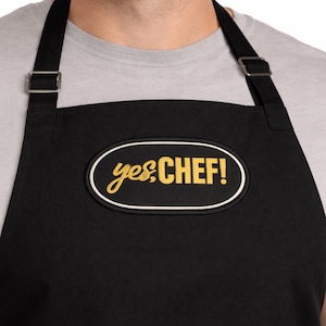 Może przedstawiać: Czarny fartuch z napisem "yes, CHEF!" w żółtym kolorze w owalu z białym obramowaniem. Fartuch posiada regulowane paski z metalowymi klamrami.