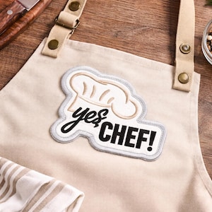 May include: Beige apron with a chef hat patch that says "yes CHEF!" in black script. The apron has adjustable straps and is made of a sturdy fabric. A striped towel is visible at the bottom. The background is a wooden surface with tomatoes and a knife.