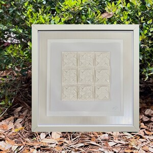 Może przedstawiać: Biała rama z obrazem przedstawiającym geometryczny wzór wykonany z białego papieru. Obraz jest oprawiony w białą ramę z srebrną obwódką.